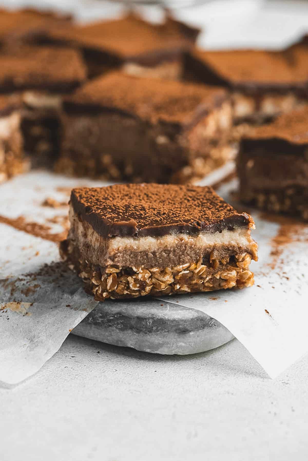 A close-up of a Tiramisu Protein Bar with three layers: an oat base, a creamy middle, and a chocolate top dusted with cocoa powder, resting on parchment paper with more protein bars in the background.