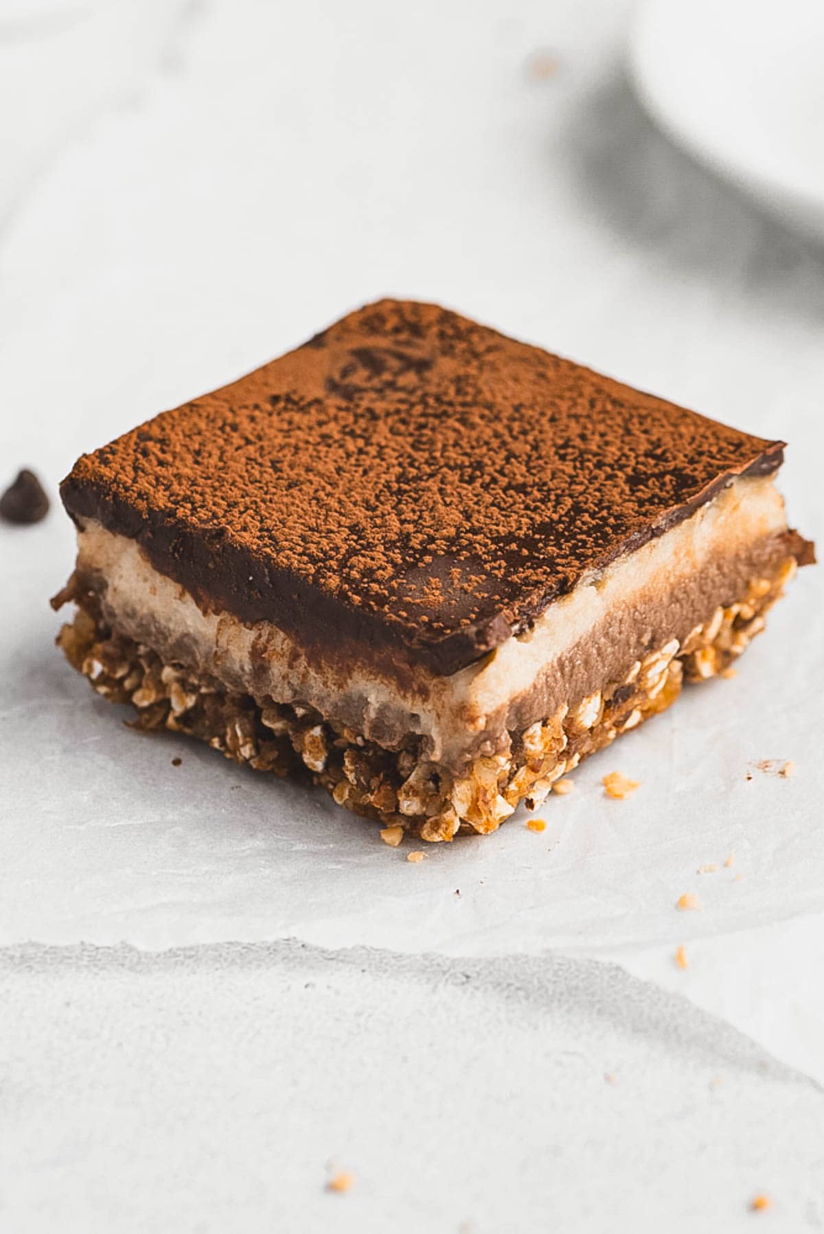 A close-up of Tiramisu Protein Bars with a granola base, creamy middle layer, and a chocolate top dusted with cocoa powder, placed on a light-colored surface.