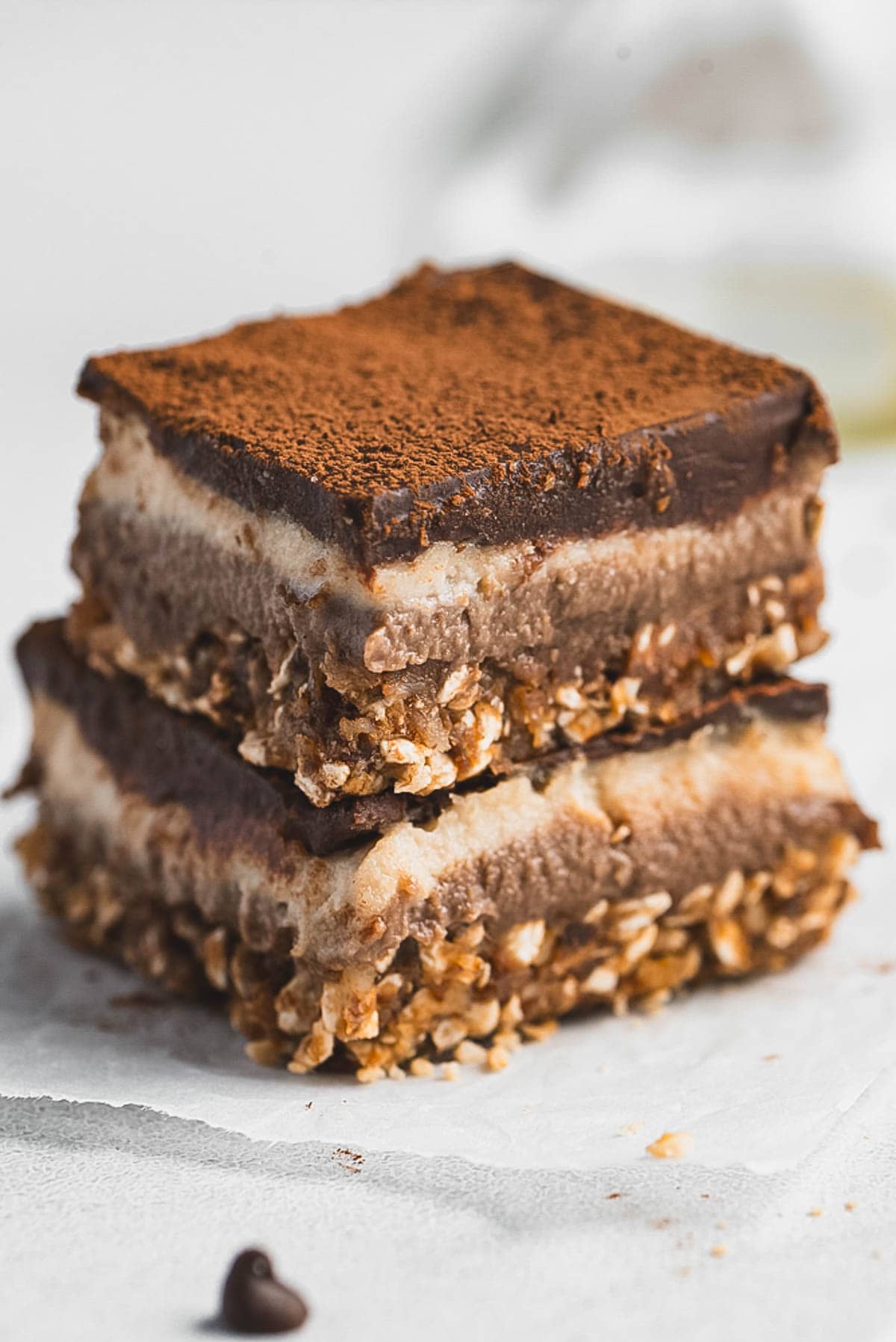 Two stacked Tiramisu Protein Bars with a crumbly oat base, creamy middle layer, and a top layer dusted with cocoa powder rest on parchment paper. A chocolate chip is visible nearby.
