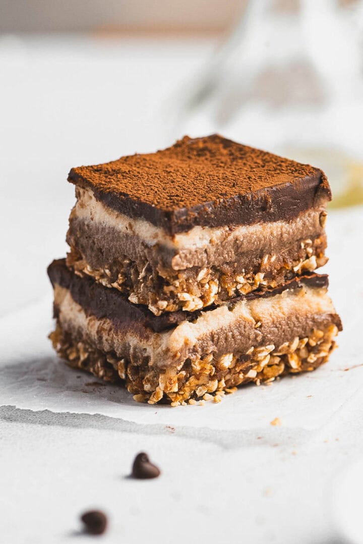 Two stacked Tiramisu Protein Bars with a crumbly oat base, creamy layered middle, and a dusting of cocoa powder on top, sitting on a white surface with a few chocolate chips nearby.