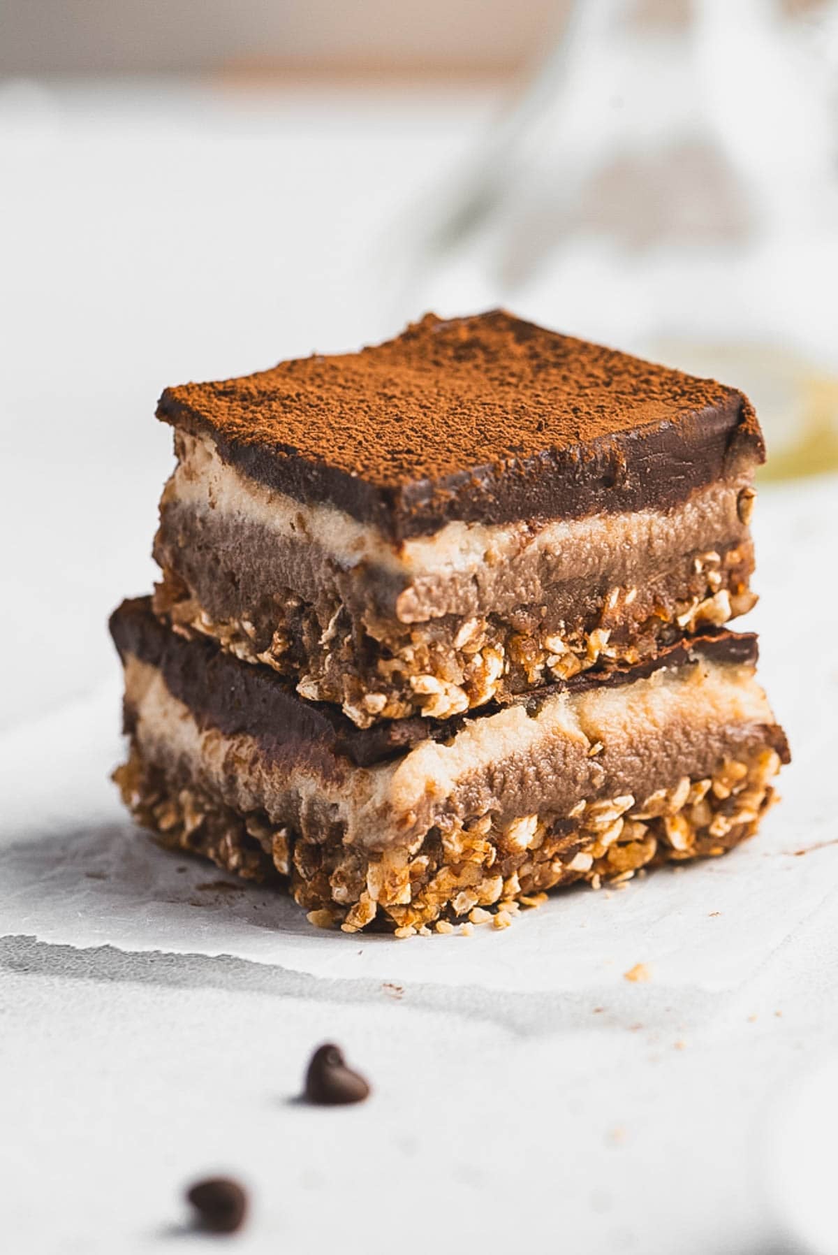 Two stacked Tiramisu Protein Bars with a crumbly oat base, creamy layered middle, and a dusting of cocoa powder on top, sitting on a white surface with a few chocolate chips nearby.