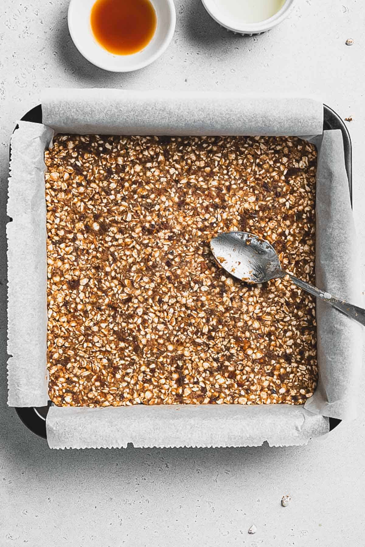 A square baking pan lined with parchment paper is filled with an even layer of unbaked Tiramisu Protein Bars oat mixture. A spoon rests on top, while two small bowls with liquid ingredients are visible in the background.