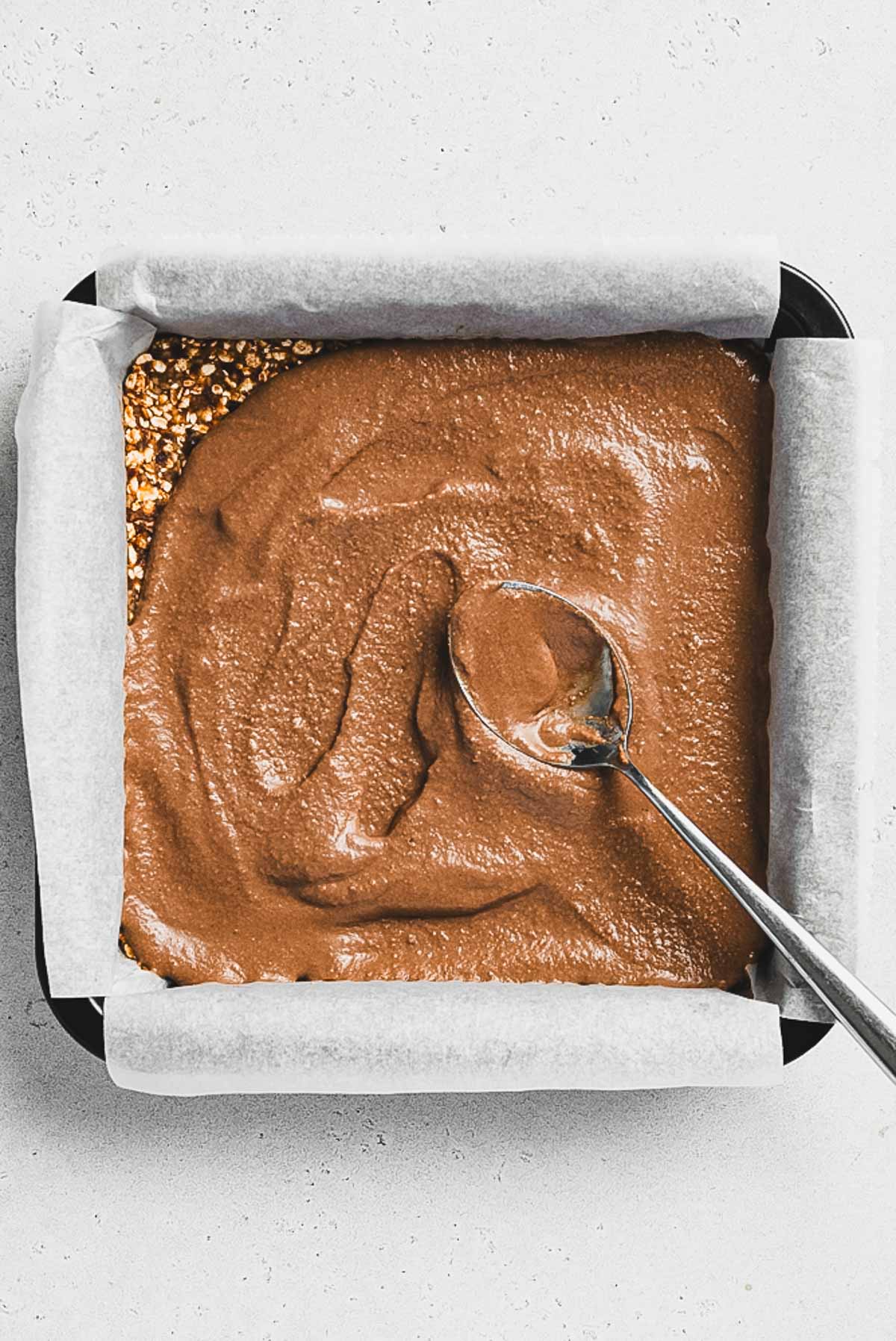 A square baking pan lined with parchment paper holds a coarse crust topped with a rich chocolate mixture, being spread with a spoon to create delicious Tiramisu Protein Bars.