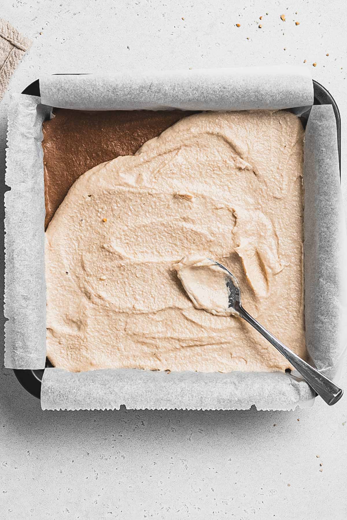 A square cake in a parchment-lined baking pan, partially covered with a layer of light brown frosting being spread with a metal spoon—these Tiramisu Protein Bars sit on a light-colored surface, ready to enjoy.