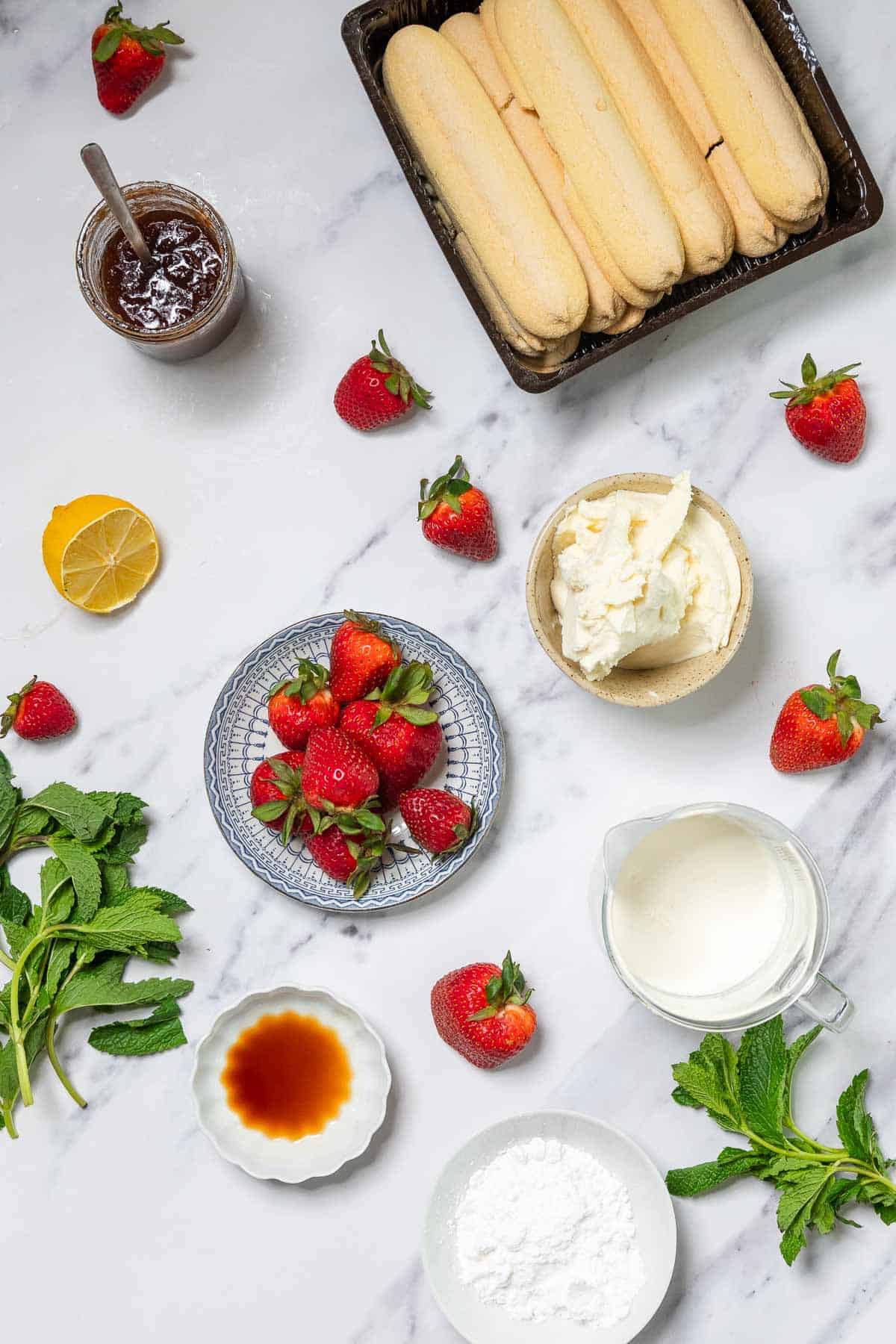 A flat lay of ingredients for a No-Bake Strawberry Tiramisu, featuring fresh strawberries, ladyfinger biscuits, mascarpone cheese, heavy cream, powdered sugar, vanilla extract, lemon half, mint leaves, and a jar of jam.