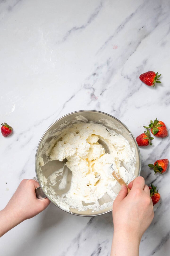 A person mixing whipped cream in a metal bowl on a marble surface, with several fresh strawberries scattered nearby&mdash;perfect for preparing a No-Bake Strawberry Tiramisu.