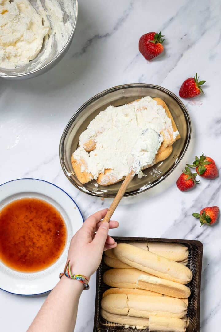 A hand spreads cream over ladyfinger biscuits in a glass dish, preparing No-Bake Strawberry Tiramisu. Nearby are a bowl of cream, strawberry syrup, fresh strawberries, and more ladyfingers on a white marble surface.