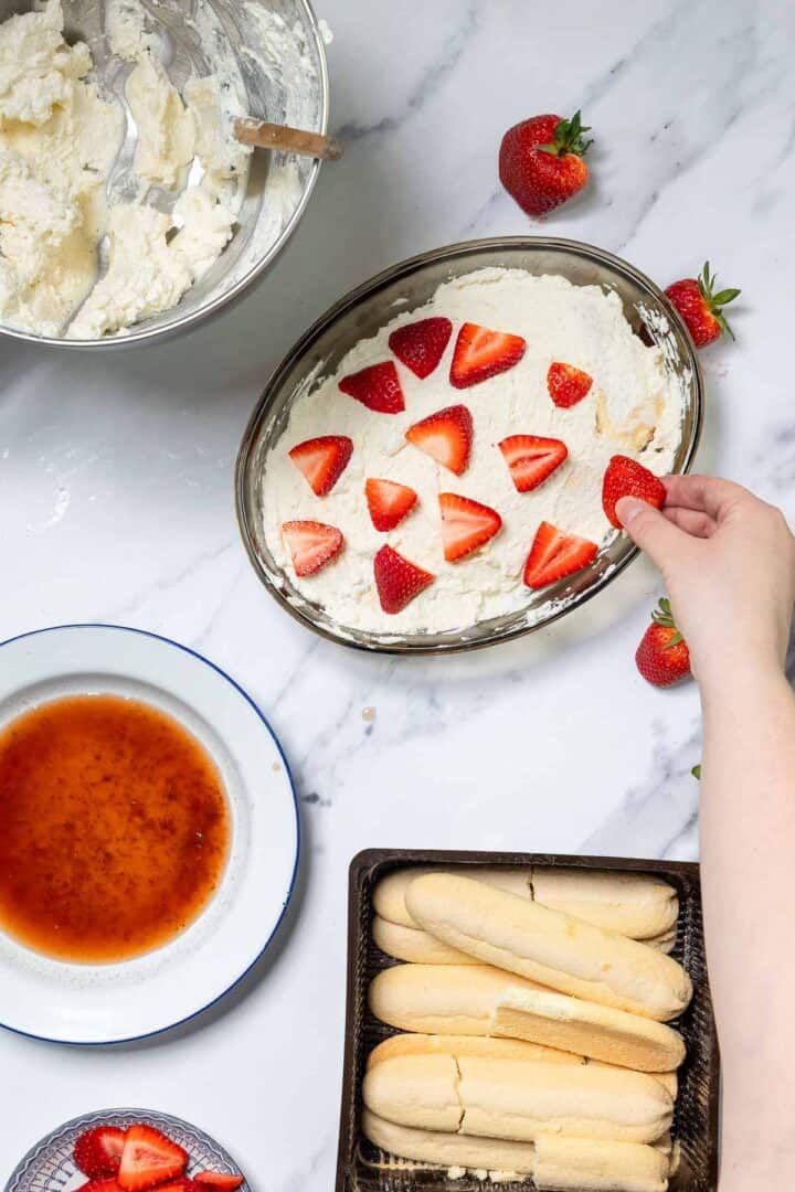 A person arranges sliced strawberries on top of a No-Bake Strawberry Tiramisu in a glass dish. Surrounding the dessert are a bowl of whipped cream, a bowl of liquid, fresh strawberries, and ladyfinger cookies.