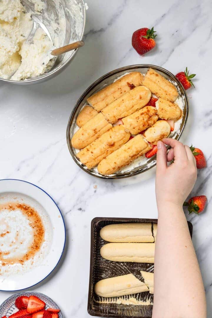 A hand assembles a No-Bake Strawberry Tiramisu, placing sliced strawberries between ladyfinger layers in an oval dish. Whipped cream, strawberry sauce, and extra ladyfingers sit nearby on a marble countertop.