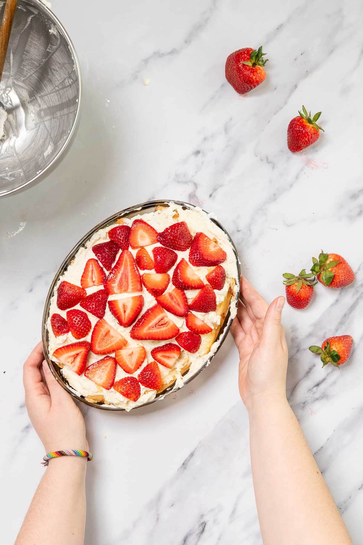 A person holding an oval dish of No-Bake Strawberry Tiramisu topped with halved strawberries on a white marble surface. A metal bowl with mixer residue and scattered strawberries are nearby.
