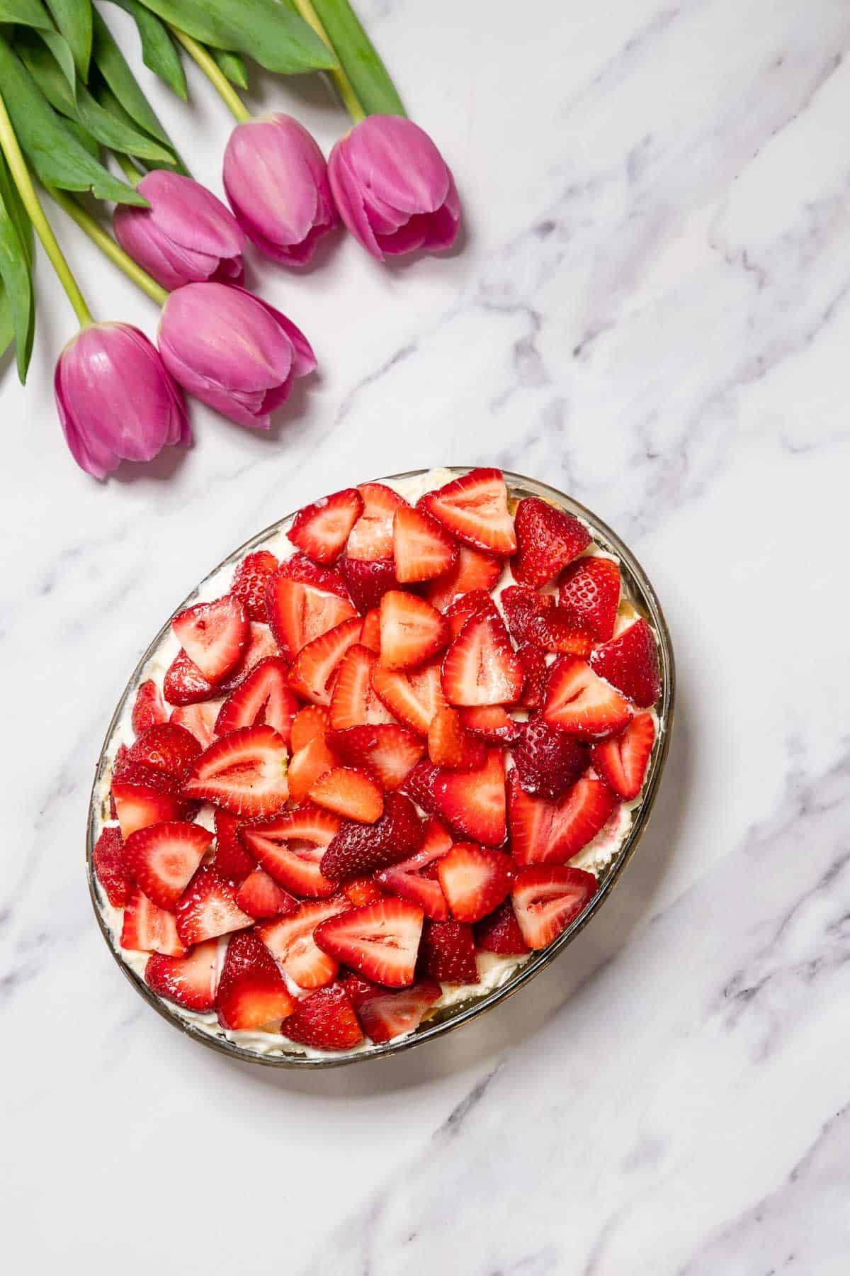 A No-Bake Strawberry Tiramisu topped with sliced strawberries sits on a marble surface next to a small bunch of pink tulips with green stems.