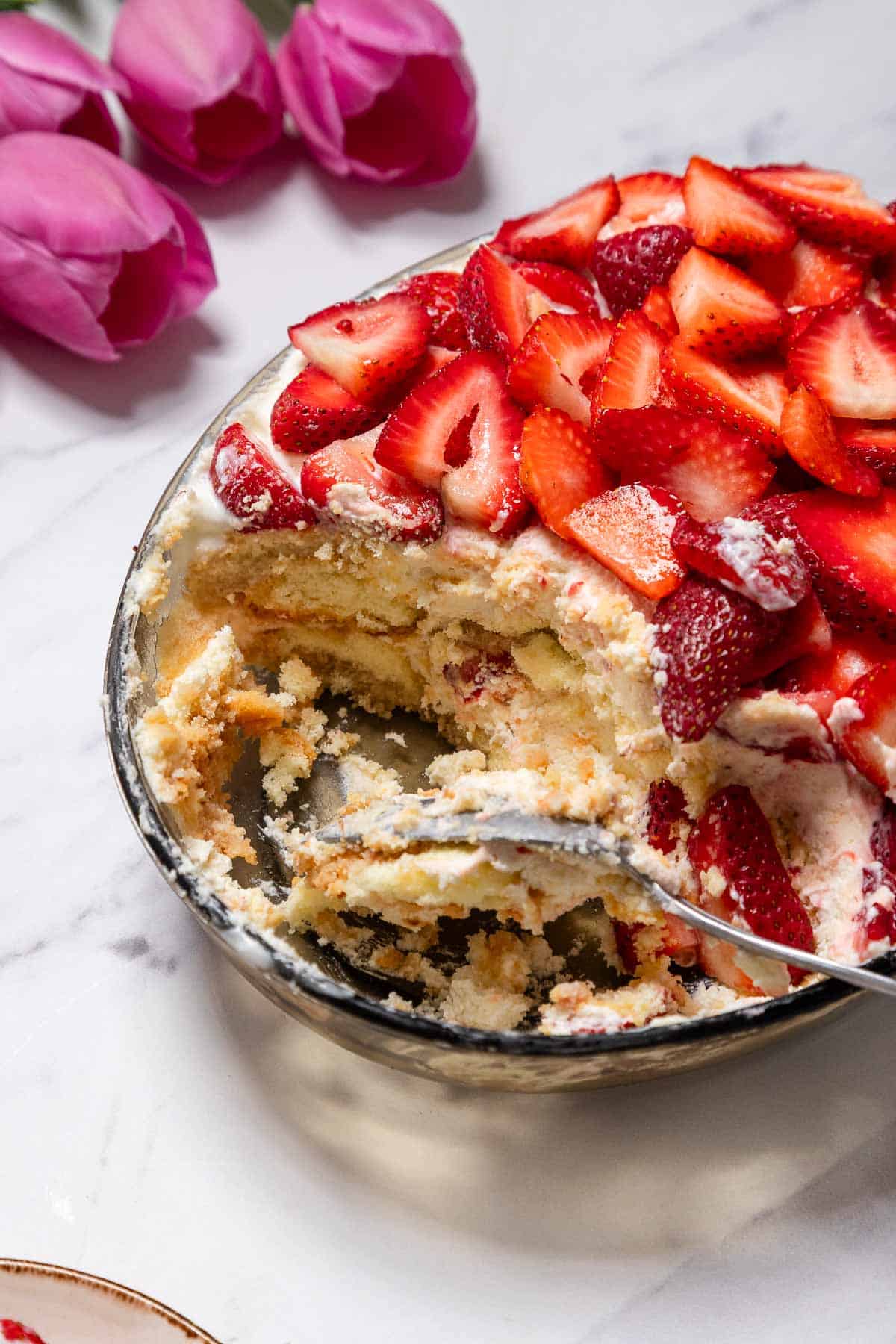 A glass dish of partially eaten No-Bake Strawberry Tiramisu with sliced strawberries on top, surrounded by crumbs and a spoon. Pink tulips are visible in the background on a marble surface.