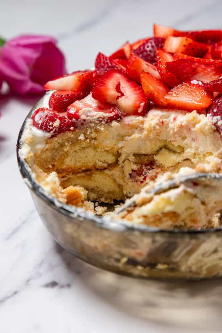 A glass bowl filled with No-Bake Strawberry Tiramisu, featuring cream, ladyfinger biscuits, and sliced strawberries on top; a portion is removed to reveal the tempting layers. A pink flower decorates the background.