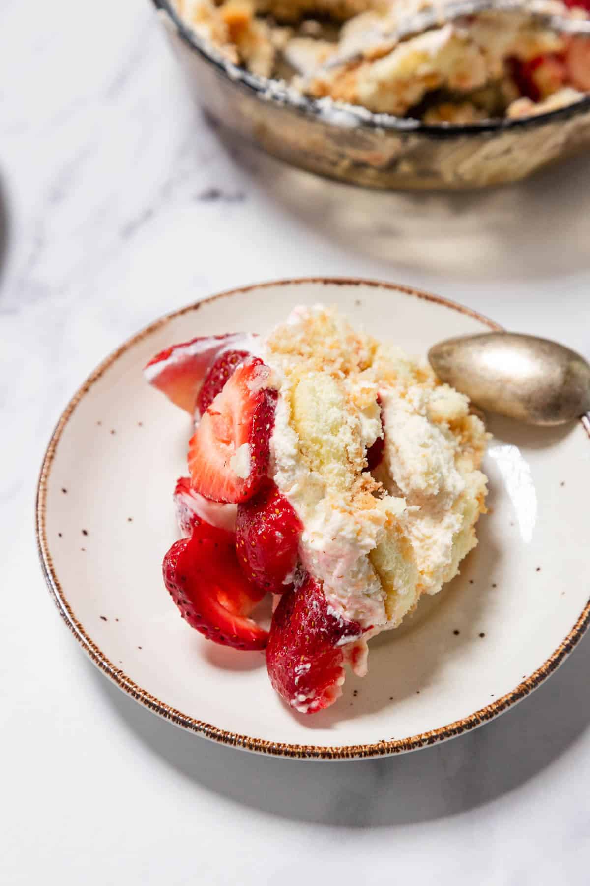 A slice of strawberry shortcake with whipped cream and fresh strawberries is served on a white plate with a brown rim, evoking the look of a No-Bake Strawberry Tiramisu. A metal spoon rests beside it, with a baking dish in the background.
