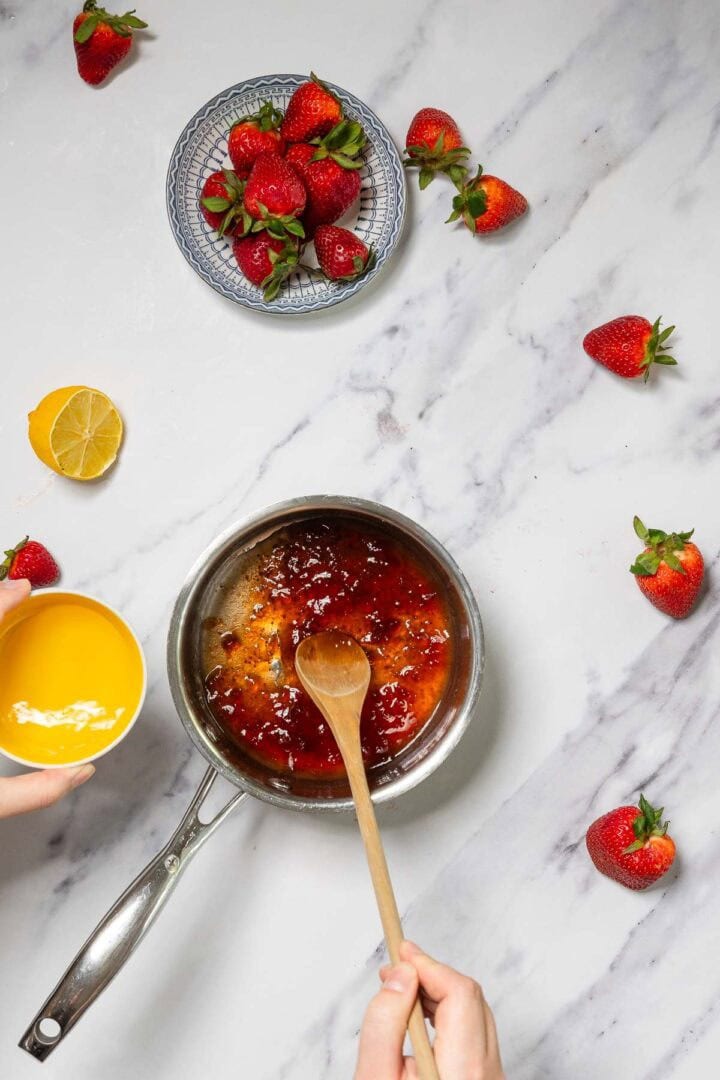 A person stirs a saucepan of strawberry sauce for No-Bake Strawberry Tiramisu with a wooden spoon, holding a bowl of yellow liquid, surrounded by fresh strawberries, a lemon half, and a small bowl of whole strawberries on a marble countertop.