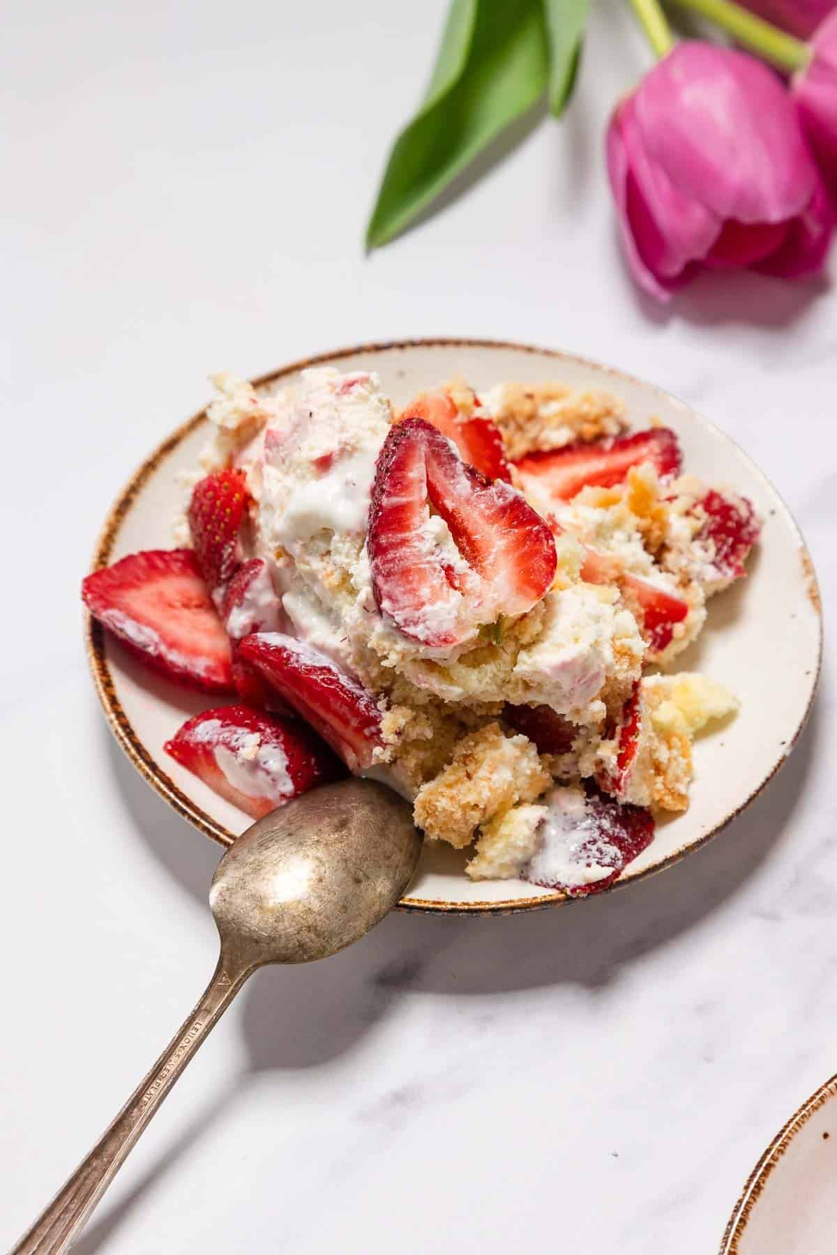 A plate with No-Bake Strawberry Tiramisu topped with sliced strawberries and whipped cream, next to an old-fashioned spoon. Pink tulips are partially visible in the background on a white surface.