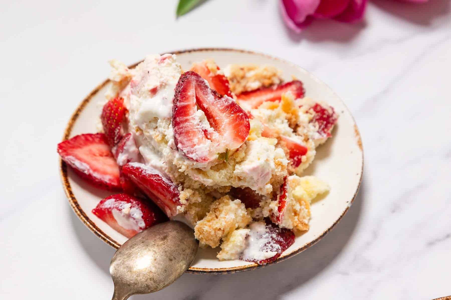 A small plate with a serving of No-Bake Strawberry Tiramisu, featuring sliced strawberries, whipped cream, and crumbled cake, with an antique-style spoon beside the dessert on a white surface.