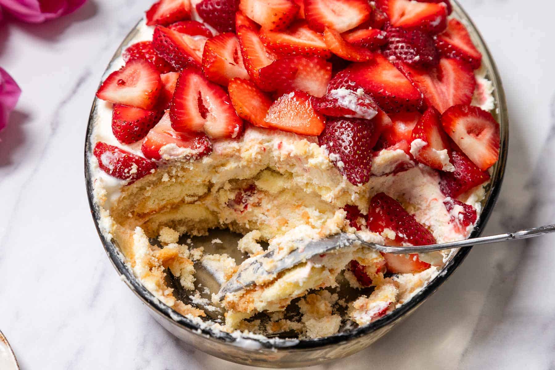 A glass dish filled with No-Bake Strawberry Tiramisu, featuring layers of whipped cream and ladyfingers, topped with sliced fresh strawberries. A serving spoon rests inside where a portion has been scooped out.