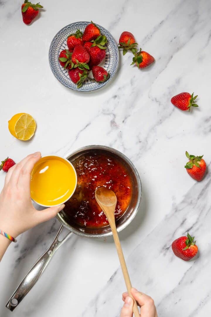 A person pours orange juice from a small bowl into a saucepan containing a red mixture for No-Bake Strawberry Tiramisu, while stirring with a wooden spoon. Fresh strawberries and a halved lemon are scattered on a marble countertop.