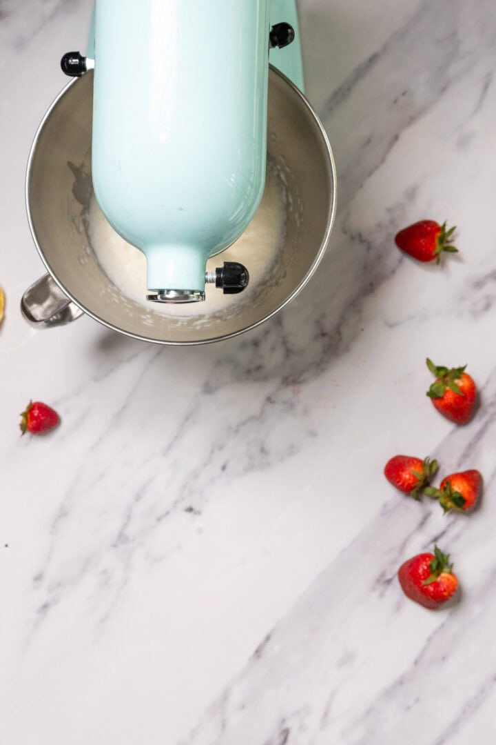 A light blue stand mixer with batter in the bowl sits on a marble countertop, ready to help whip up a No-Bake Strawberry Tiramisu. Several fresh strawberries are scattered nearby, adding a burst of color and flavor.