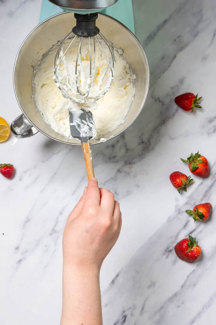 A hand holds a spatula mixing creamy batter in a stand mixer bowl, preparing for No-Bake Strawberry Tiramisu. Several strawberries and a lemon wedge are placed on the marble countertop nearby.