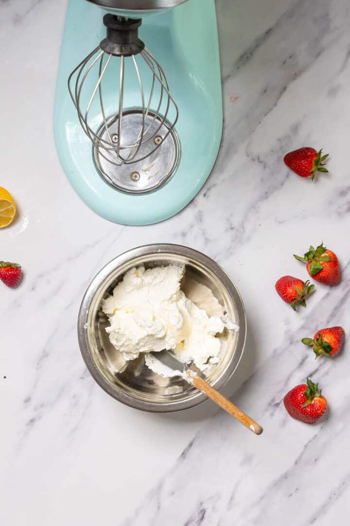 A mixing bowl with whipped cream for No-Bake Strawberry Tiramisu and a spatula sits on a marble countertop. Nearby are fresh strawberries, a lemon wedge, and a stand mixer with a whisk attachment.