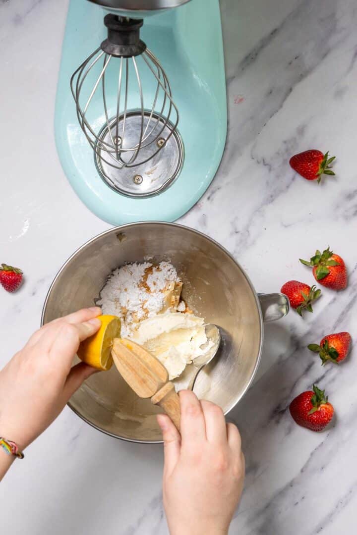 A person squeezes a lemon half over a metal mixing bowl with cream cheese, powdered sugar, and vanilla extract for a No-Bake Strawberry Tiramisu. The bowl is attached to a stand mixer, and fresh strawberries rest on the marble countertop nearby.