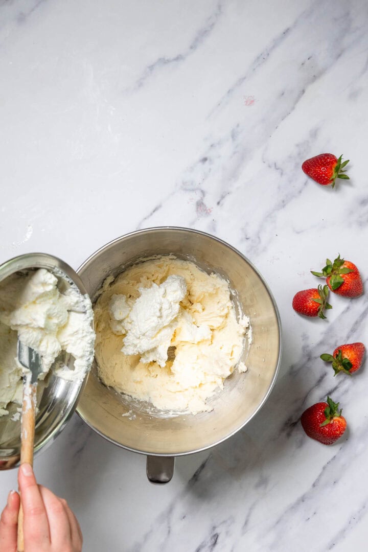 A hand uses a spatula to transfer whipped cream from a saucepan into a mixing bowl on a marble surface, preparing for No-Bake Strawberry Tiramisu, with three whole strawberries nearby.