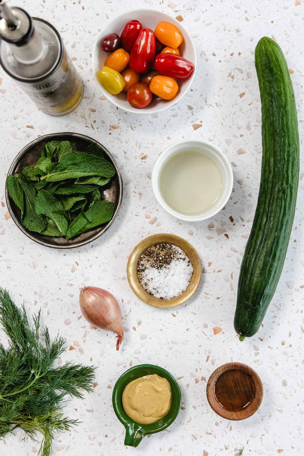 Aerial view of a countertop with ingredients for Tomato and Smashed Cucumber Salad: cucumber, cherry tomatoes, shallot, fresh mint, fresh dill, olive oil, vinegar, salt and pepper, mustard, and a small wooden bowl.