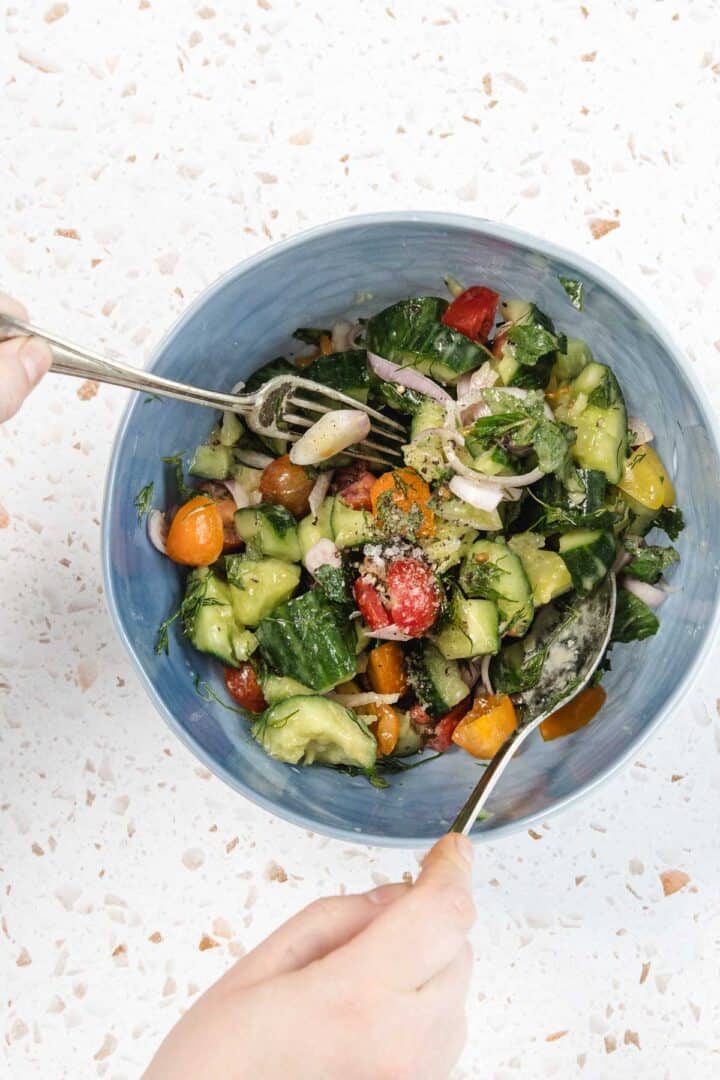 A person uses a fork and spoon to toss a Tomato and Smashed Cucumber Salad in a blue bowl. The mix of cucumbers, cherry tomatoes, red onion, greens, and herbs sits on a white speckled countertop.