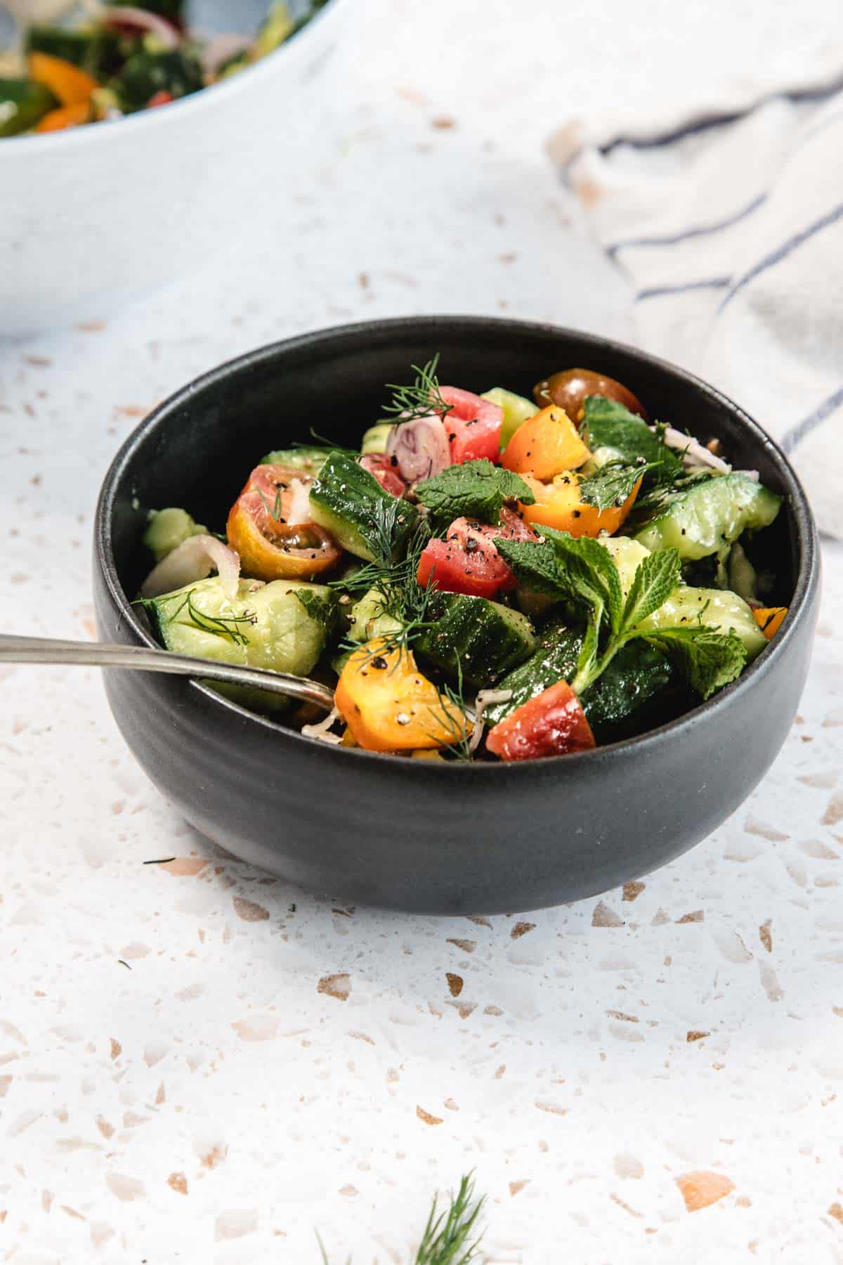 A black bowl filled with Tomato and Smashed Cucumber Salad—chopped cucumbers, tomatoes, and fresh herbs—sits on a white speckled surface with a fork and striped napkin nearby.