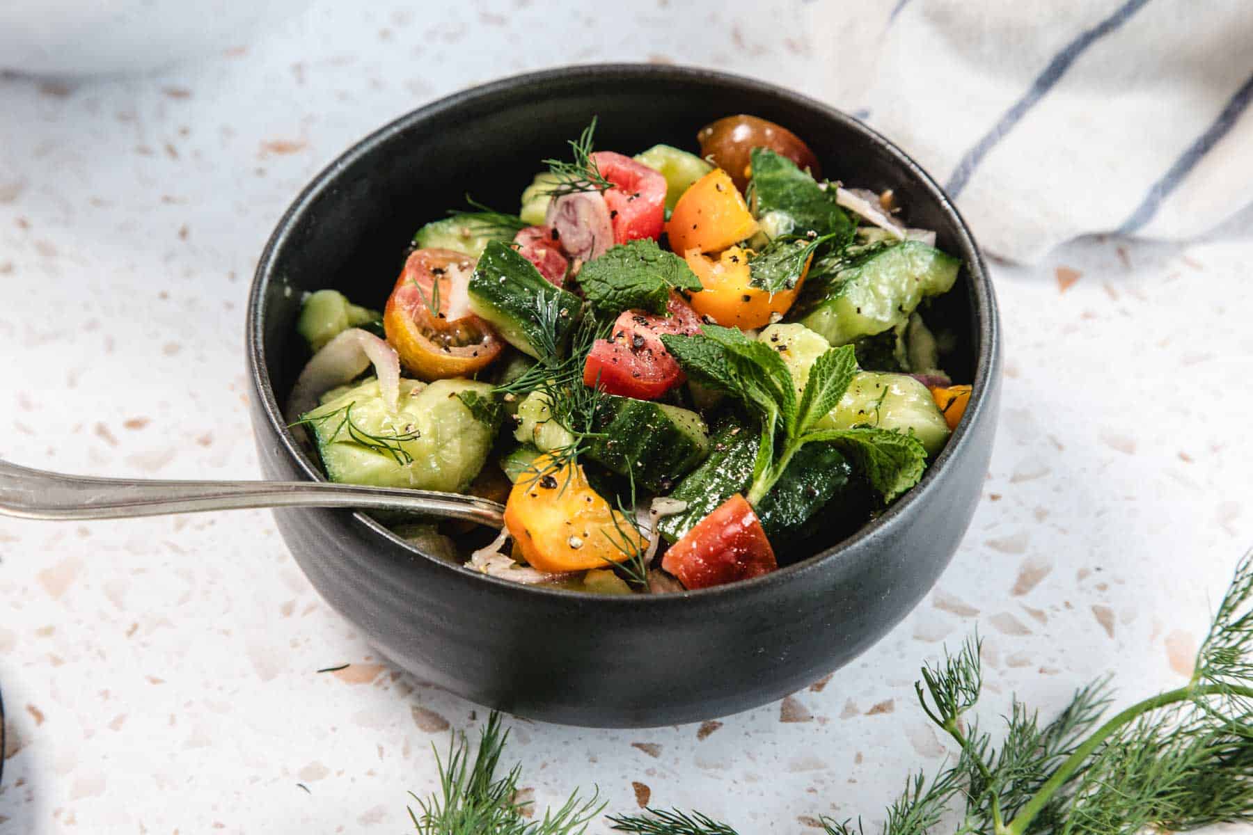 A black bowl filled with Tomato and Smashed Cucumber Salad—chopped cucumbers, assorted cherry tomatoes, red onions, and herbs. A silver spoon rests inside, with fresh dill and a striped cloth nearby.