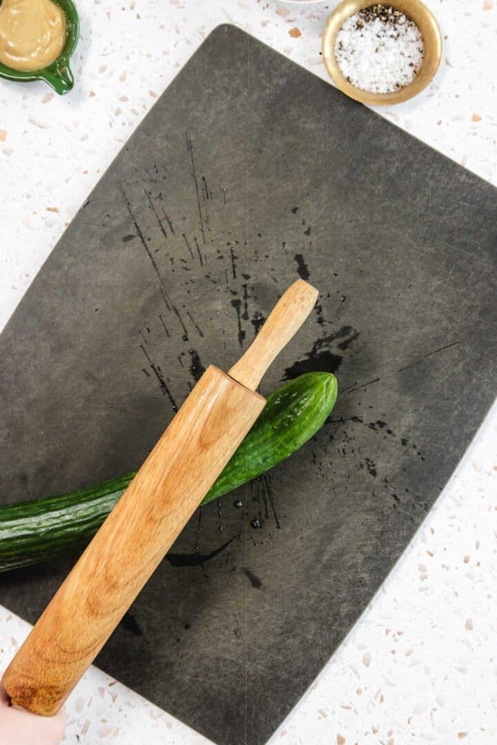 A hand uses a wooden rolling pin to smash a cucumber on a black cutting board, prepping ingredients for Tomato and Smashed Cucumber Salad. A small bowl of salt and a green dish with sauce are nearby on a white countertop.