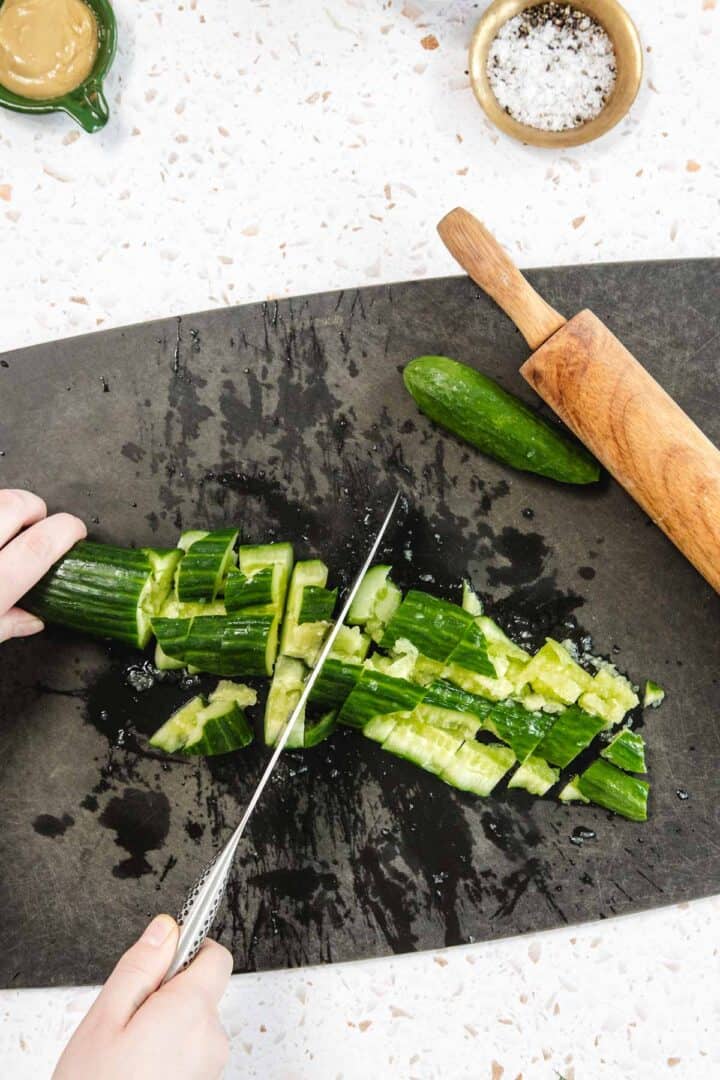 A person chops smashed cucumber pieces for a Tomato and Smashed Cucumber Salad with a knife on a black cutting board. A wooden rolling pin, whole cucumber, salt, and other ingredients are nearby on a light countertop.