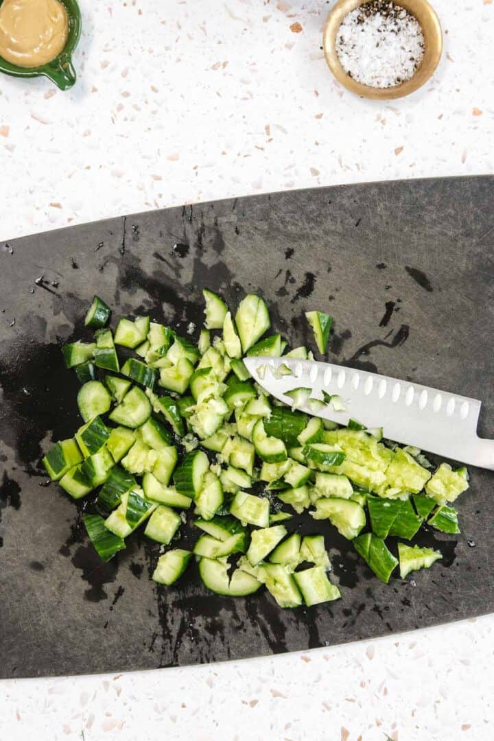 A knife and roughly chopped cucumber pieces, perfect for a Tomato and Smashed Cucumber Salad, are on a dark cutting board. A small bowl of salt and a dish of mustard rest on a white countertop nearby.