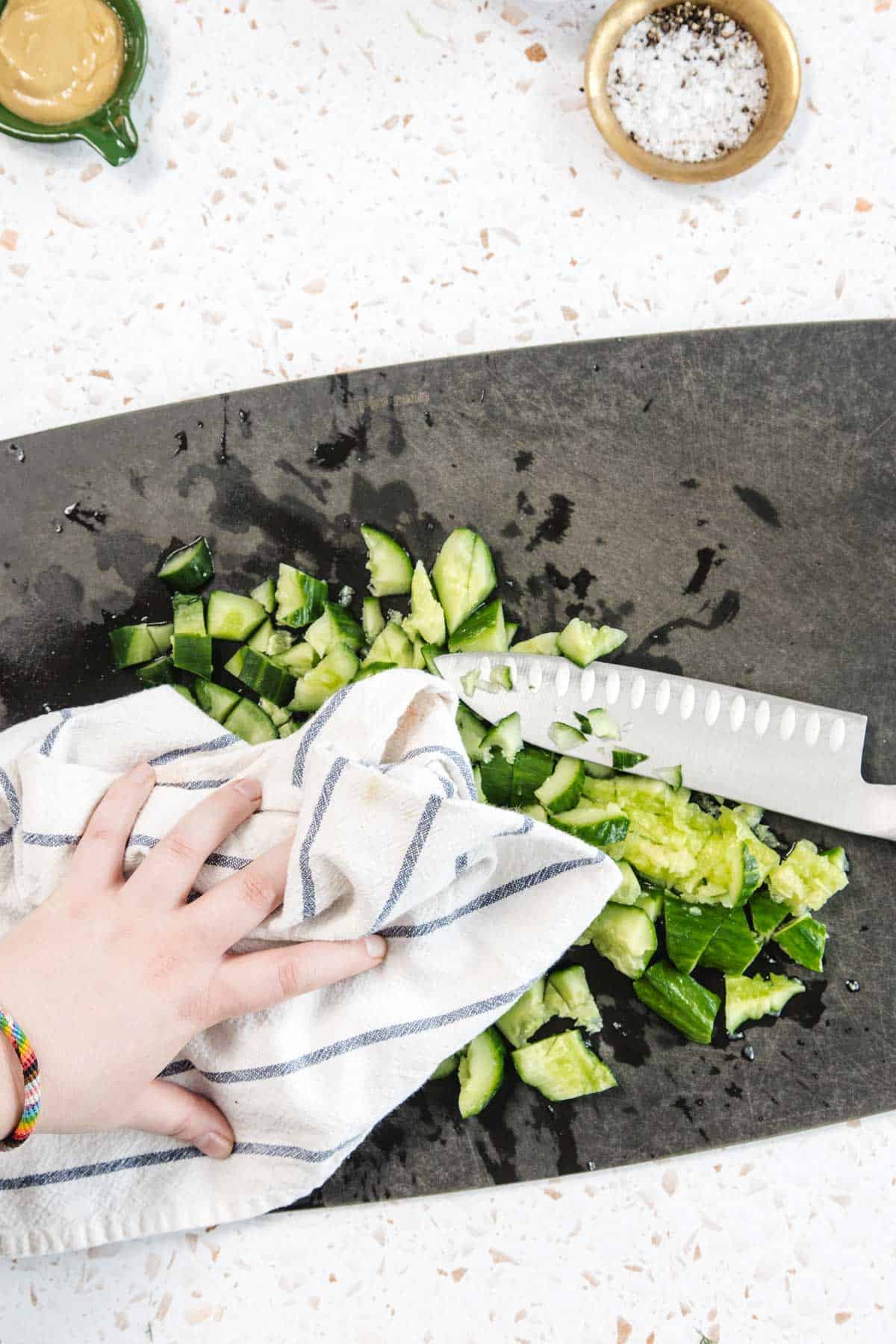 A hand holding a striped kitchen towel crushes chopped cucumber pieces for a Tomato and Smashed Cucumber Salad on a black cutting board, with a knife and small bowls of salt and mustard visible nearby.