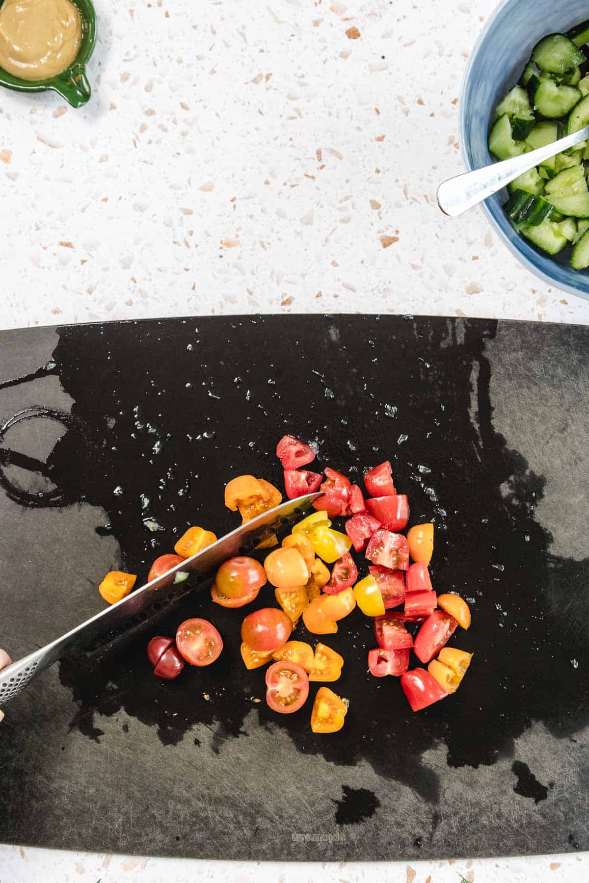 A knife slices through assorted red and yellow cherry tomatoes for a Tomato and Smashed Cucumber Salad on a black cutting board, with a blue bowl of cucumbers and a juicer with a wedge of lime nearby on a speckled countertop.