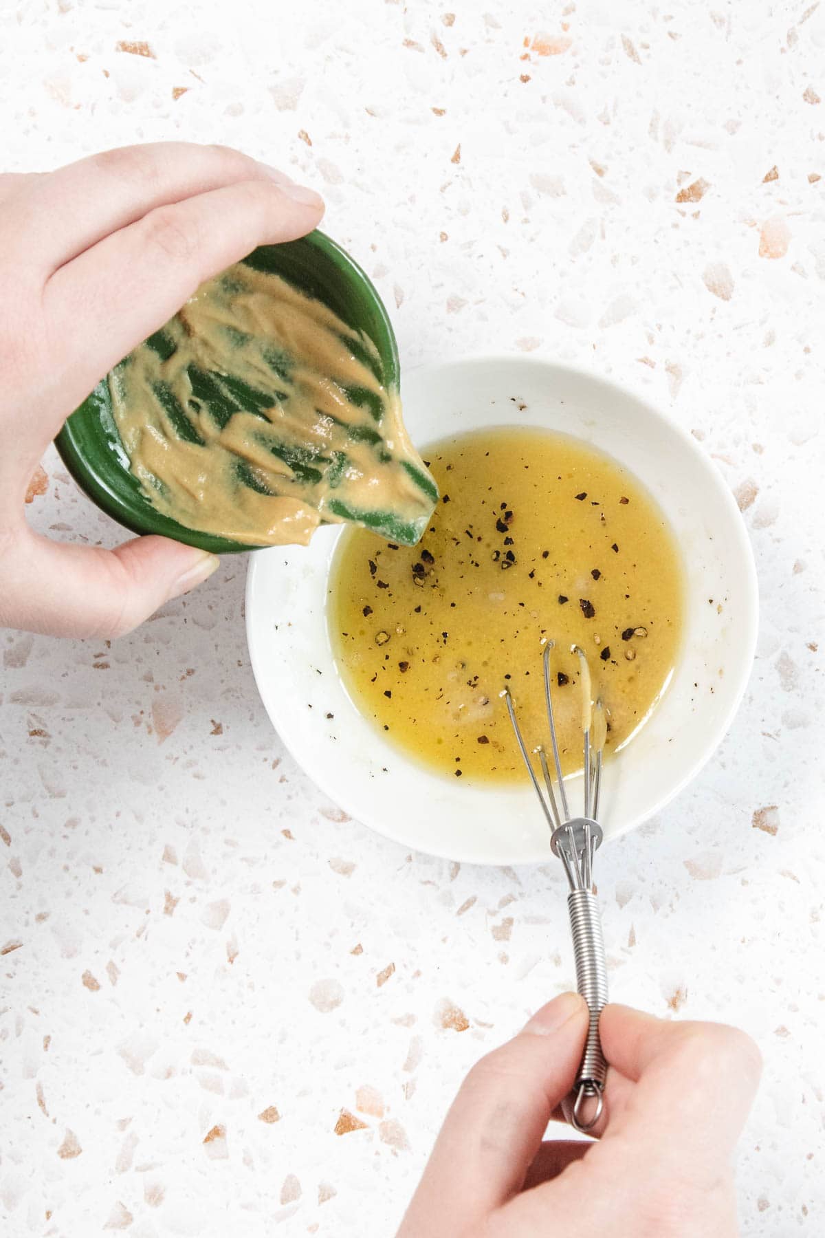 A person prepares a Tomato and Smashed Cucumber Salad by pouring mustard from a small green bowl into a white bowl with oil, vinegar, and cracked black pepper, while holding a small whisk ready to mix the ingredients.