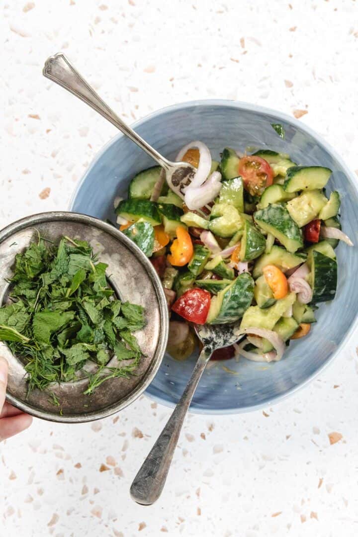 A hand holds a small metal bowl of chopped fresh herbs above a blue bowl containing a Tomato and Smashed Cucumber Salad with onions, served with two utensils on a speckled countertop.
