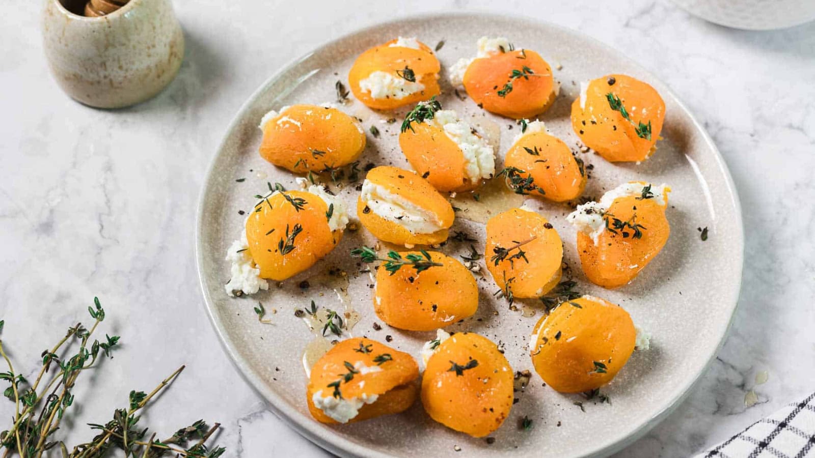 A plate with halved dried apricots stuffed with white cheese and garnished with herbs, placed on a light marble surface with a sprig of fresh thyme nearby.