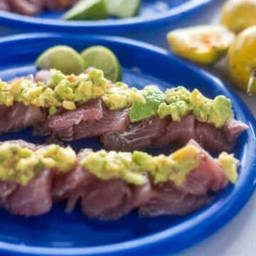 Slices of raw fish topped with diced avocado are arranged on a blue plate, with slices of lime and whole passion fruits visible in the background.