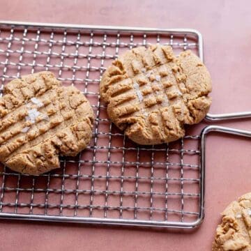 Four peanut butter cookies with crisscross fork marks are on a metal cooling rack, with another cookie and a spoonful of peanut butter on a pink surface nearby.