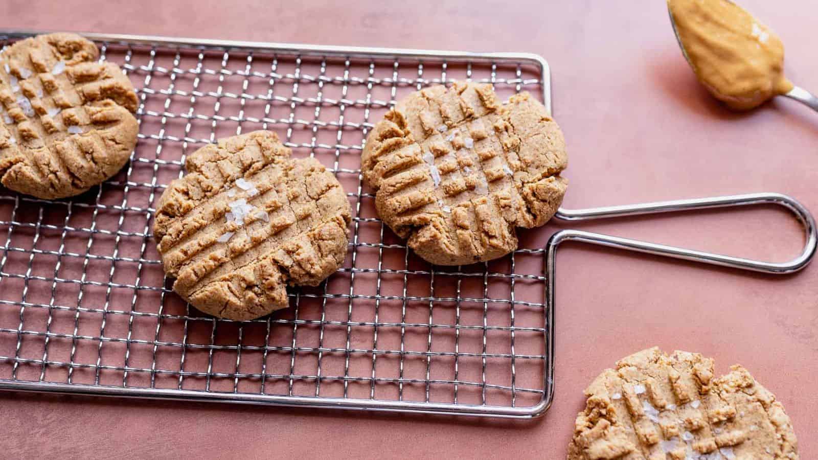 Four peanut butter cookies with crisscross fork marks are on a metal cooling rack, with another cookie and a spoonful of peanut butter on a pink surface nearby.