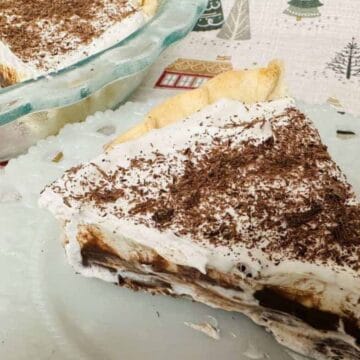 A slice of chocolate pudding pie topped with whipped cream and chocolate shavings sits on a white plate. The rest of the pie, with one slice missing, is in a glass pie dish in the background.