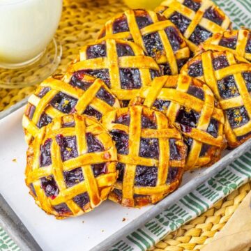 A tray of small lattice-topped blueberry pies is placed on a striped cloth. Next to it are a glass of milk, a halved orange, and a wooden spoon filled with fresh blueberries.