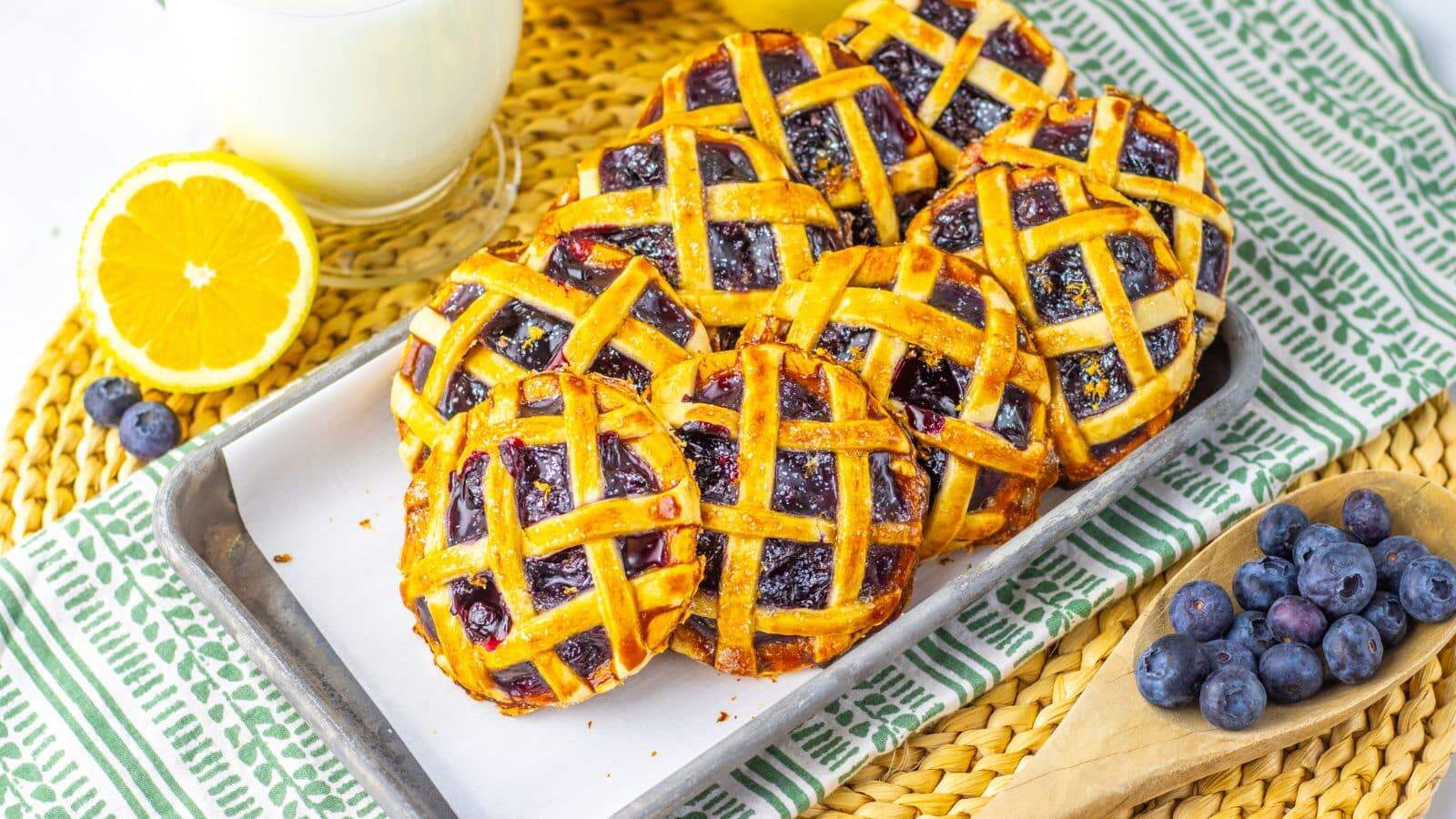 Seven blueberry pies with lattice tops on a tray, accompanied by fresh blueberries, half an orange, and a glass of milk on a woven mat.