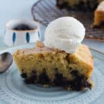 A slice of Blueberry Polenta Cake with visible dark berries is topped with a scoop of vanilla ice cream on a patterned plate. A spoon and small bowl sit nearby, with a cooling rack and more cake in the background.