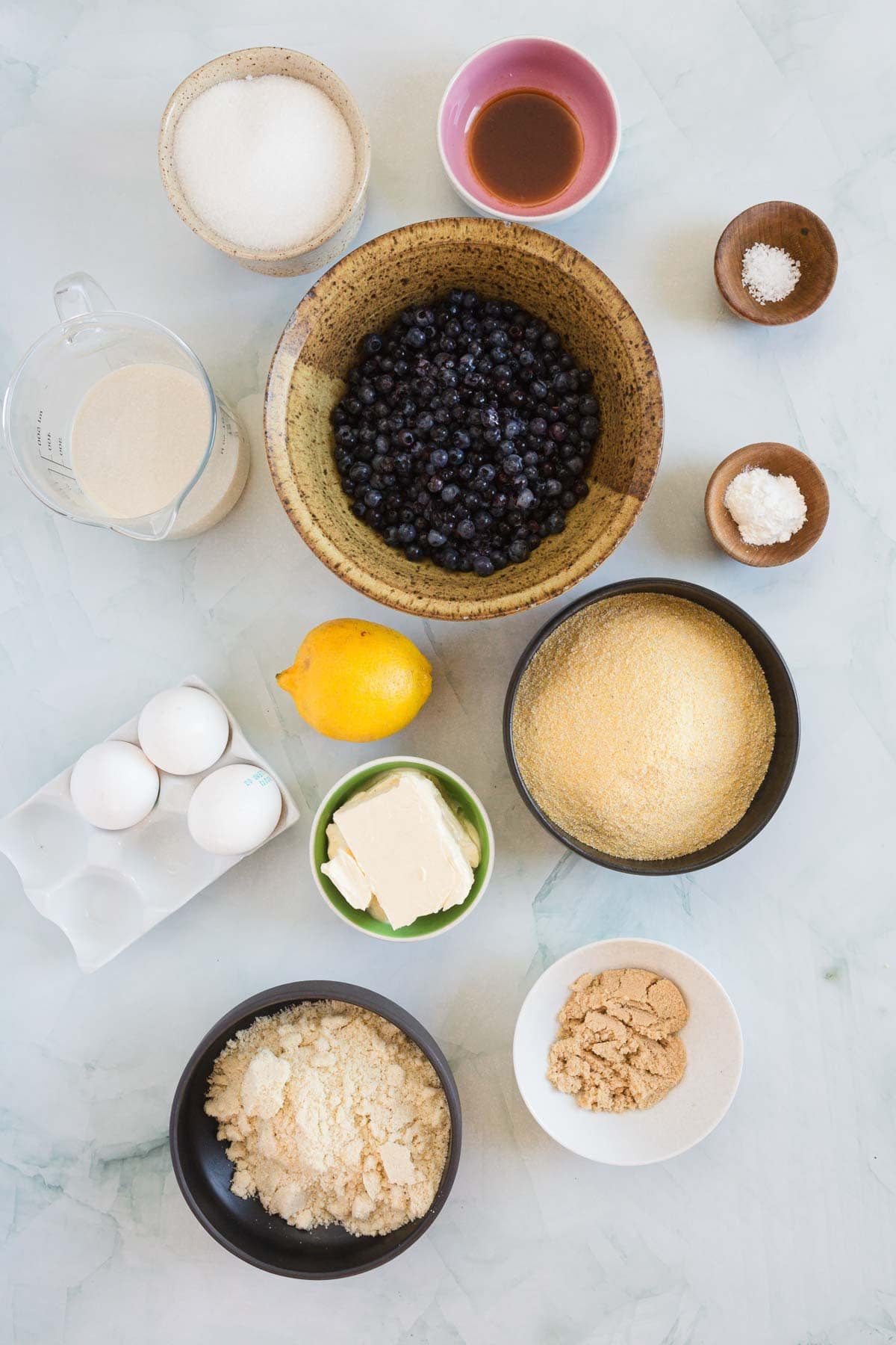 A flat lay of baking ingredients for Blueberry Polenta Cake on a marble surface, including blueberries, eggs, lemon, butter, sugar, brown sugar, cornmeal, flour, almond flour, vanilla extract, baking powder, and salt in bowls and cups.
