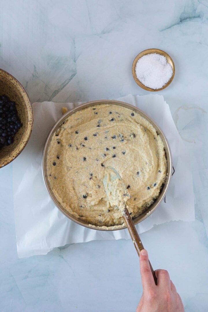 A hand is stirring Blueberry Polenta Cake batter with blueberries using a spatula. The bowl sits on parchment paper atop a light countertop. Nearby are fresh blueberries and a small dish of sugar, ready to add extra sweetness.