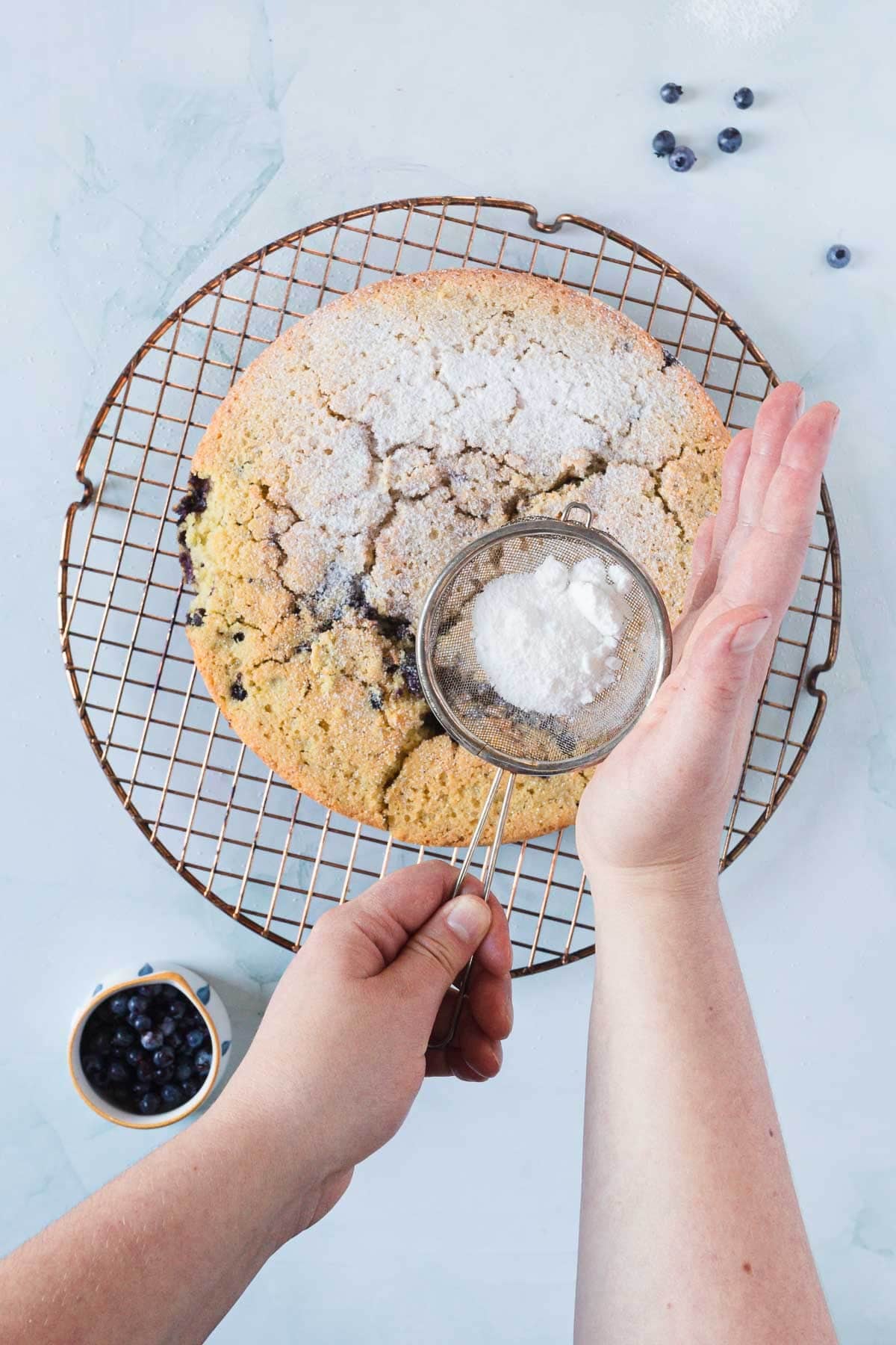 A person dusts powdered sugar over a round Blueberry Polenta Cake on a wire cooling rack, using a small sieve. A bowl of blueberries and a few loose berries rest nearby on a light-colored surface.
